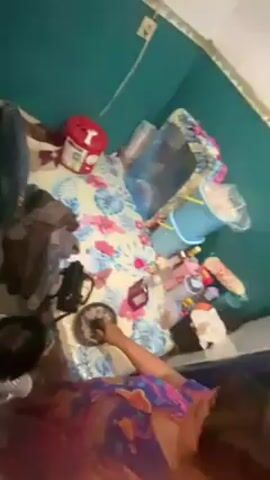 From a high angle, a person is grinding something on a plate using a tool. The person is standing near a bed covered with a floral patterned sheet. There are various items scattered on the bed, including containers, a small bucket, and other miscellaneous objects. A second person, a small child, is visible at the bottom of the frame. The walls are painted teal.