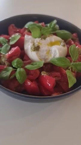 A close-up shot of a dark blue bowl filled with a tomato and burrata salad. The tomatoes are chopped and arranged in the bowl, topped with a ball of burrata cheese. The burrata is drizzled with olive oil and pesto, and garnished with fresh basil leaves. The salad is placed on a white surface. The video is shot from a slightly elevated angle and the focus is on the salad.