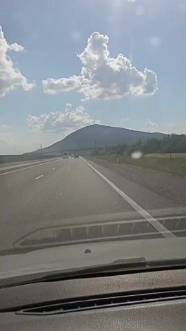 A perspective from inside a vehicle shows a highway with light traffic leading towards a large, gently sloping mountain under a partly cloudy sky. The sun is visible and casting a bright glare. The road has clear white lane markings and is bordered by a grassy shoulder. A bridge or overpass is visible in the distance to the left. The dashboard of the vehicle is visible in the foreground.