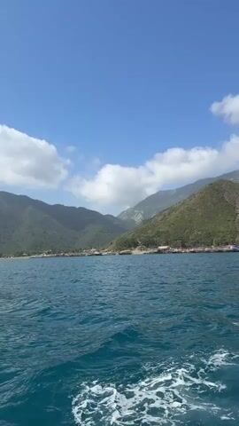 The video shows a view of a calm, blue sea, from the surface, likely taken from a boat moving across the water. The surface is slightly choppy. In the distance, there's a coastline with green, mountainous terrain. Some buildings or structures are visible along the coastline, appearing small and indistinct due to the distance. The sky is a light blue with scattered white clouds.