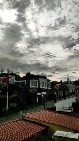 The camera captures a still, wide-angle shot of a suburban neighborhood under a dramatic sky. The sky is heavily overcast with a mixture of gray and white clouds, allowing patches of blue sky to peek through. The sun's light filters through the clouds, creating a diffused illumination over the scene.

In the foreground, the red tile roofs of several houses are visible, suggesting a residential area. A road winds through the neighborhood, lined with houses of varying designs. Some houses are multi-story with modern architectural elements, including large windows and neutral color palettes. Other houses are smaller and more traditional in style, with white walls and darker roofs. 

Green trees and vegetation are interspersed among the houses, adding a touch of nature to the urban landscape. In the distance, a tall, slender tower is visible, possibly a telecommunications or water tower, providing a focal point on the horizon. The overall impression is that of a quiet, upscale neighborhood under a cloudy sky.