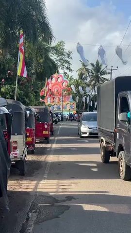 The video shows a street in Sri Lanka during a Buddhist festival or celebration.  A large, colorful, lighted religious display with multiple tiers is centered on a roundabout or in the middle of the road. The display is adorned with images. Buddhist flags are erected to the left and right of the display. Numerous white lanterns hang overhead, strung across the road. Several parked tuk-tuks are visible on the side of the road, along with motorcycles and a few cars. A silver car with a Sri Lankan license plate drives on the right-hand side of the road. Pedestrians are milling around the display. Palm trees and other greenery are visible in the background. The sky is partly cloudy.