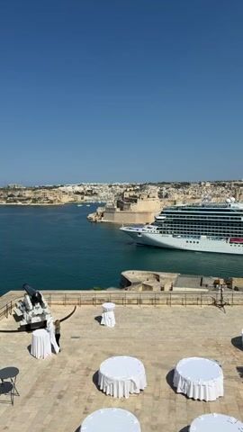 This media file captures a scenic view of a harbor in Valletta, Malta, on a clear, sunny day, suggesting it's midday or early afternoon. The foreground features a paved terrace area with several round tables covered in white tablecloths, some of which appear to be set up for an event, possibly an outdoor reception or wedding. A person, dressed in dark clothing, is adjusting the tablecloth on one of the tables. To the left, a historical cannon is mounted on a stone platform, adding to the historical ambiance of the location.

Beyond the terrace, a vast expanse of turquoise water fills the middle ground. A large, white cruise ship, identified by the visible text "Allura" on its hull, is navigating through the harbor. The ship is positioned to the right of the frame, moving from left to right. In the background, the cityscape of Valletta unfolds, characterized by its distinctive honey-colored buildings, fortified walls, and a dense urban landscape. The clear blue sky stretches across the top of the frame, indicating fair weather.

The scene evokes a sense of tranquility and grandeur, with the juxtaposition of historical elements (cannon, fortifications) and modern amenities (cruise ship, event setup). The bright sunlight and the calm waters contribute to a feeling of pleasantness and opportunity for outdoor activities. The overall impression is one of a well-preserved historical site offering a beautiful vantage point for enjoying the harbor activity.