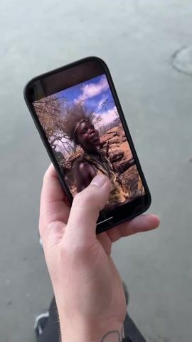 The video begins with a close-up shot of a person's hand holding a black smartphone. The phone's screen displays a video of an individual with dark skin, wearing what appears to be traditional attire made of animal hide or fur, and some necklaces. This person has distinct spiky hair, is smiling, and looks towards the camera. The background of the video on the phone shows dry, rocky terrain and sparse trees under a partly cloudy sky. A tattoo is partially visible on the wrist of the hand holding the phone.

As the video progresses, the hand and phone move out of the frame, revealing a wider shot of an urban environment. A multi-lane highway or large road is visible in the foreground, with grey asphalt and some yellow road markings. A concrete barrier separates the road from the background. In the distance, several tall buildings are under construction, with cranes visible. A blue semi-truck is parked or moving slowly behind the barrier. The sky is bright blue with scattered white clouds.

The camera then pans slightly to the left, showing more of the highway. A tall streetlight with multiple lamps stands by the road. More buildings are visible in the background, including a commercial building with large windows and text that includes "КОЛЕСО" (Koleso) and "СЕРВИС" (Service), suggesting a car service center or dealership. Another section of a building has the word "МАНСТАР" (Manstar) visible. Several cars are visible on what appears to be a parallel road or parking area behind the barrier.

Finally, the camera continues to pan, focusing on a large white semi-truck with a long trailer driving on the main road from right to left across the frame. The side of the trailer is prominently branded with "KNAUF ROTBAND®" in large, bold red letters against a dirty white background. The blue sky remains clear.