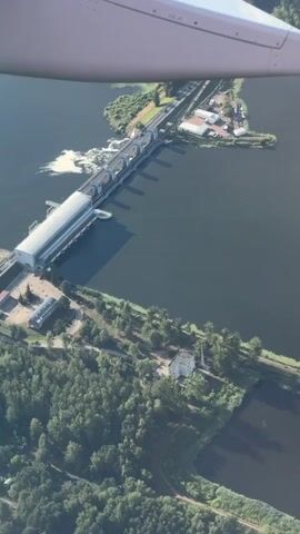 An aerial view captures a dam with a long, rectangular building running across it, possibly a hydroelectric power station. Water flows over a spillway on the left side of the dam, creating white rapids. A road and railway tracks run parallel to the dam, connecting to buildings on the shore. The surrounding landscape is lush and green, with dense forests bordering the dark, calm water. The top portion of the image shows a white airplane wing, indicating the perspective is from an aircraft. The scene is bathed in daylight, suggesting it is daytime.