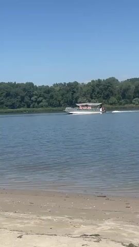 A white boat with a red and white striped canopy travels across a blue body of water. The boat is moving from left to right, and its wake is visible. A line of lush green trees forms the background. In the foreground, a sandy beach is visible. The sky is a clear, bright blue.