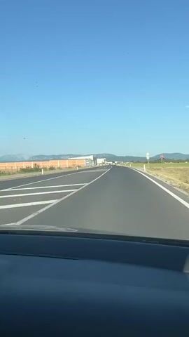 The video is shot from the perspective of a passenger inside a car, looking out the windshield and dashboard. The car is driving on a paved road under a clear blue sky. On the left side of the road, there is a long, orange-colored barrier or wall, with some green vegetation in front of it. In the distance, there appear to be some buildings and possibly mountains. The dashboard of the car is visible, with a steering wheel on the left. There are several air fresheners attached to the air vents. The road has white lane markings. The video shows the car moving forward, indicating it is in motion.
