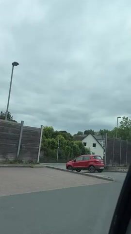 A red SUV is driving on a street under a cloudy sky. The vehicle is captured from behind and to the side. The interior of the vehicle is visible, showing a person's head and shoulders, and the back of a headrest. The surroundings appear to be suburban, with trees, houses, and sound barriers along the road. The video is short and does not show any significant actions or interactions.