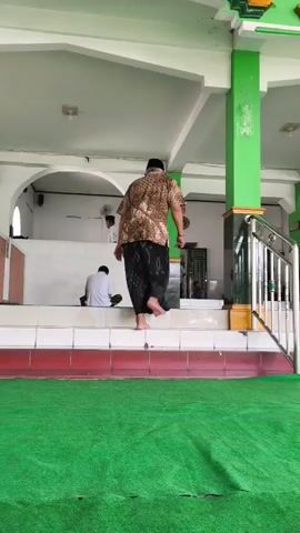 A man wearing a patterned shirt and a sarong walks up a set of stairs in a mosque. Another man in a white shirt is sitting on the stairs. The mosque has green carpet on the floor and white walls with green accents.