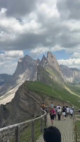 This media file captures a scenic view of a mountain range, likely in the Dolomites, Italy, based on the distinctive jagged peaks. The foreground features a wooden railing and a paved walkway populated by several people. They appear to be tourists, dressed in casual summer attire, observing the breathtaking landscape. The walkway follows a path along a steep grassy slope, with a dramatic drop-off to the left, revealing layered rock formations. In the background, a grand mountain peak with multiple sharp summits dominates the horizon. The sky is a mix of blue and dramatic, grey-tinged clouds, suggesting a dynamic weather system possibly experienced earlier or expected later in the day. The lighting indicates it's likely daytime, possibly late morning or early afternoon. The mood appears relaxed and awe-inspired, as people take in the natural beauty. No text is visible in the media file.
