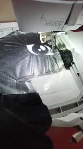 The video shows an embroidery machine in operation. The needle is down, sewing through a black fabric layered with a shiny material, possibly plastic or vinyl, and a white stabilizer. The embroidery hoop is securely holding the fabric. The machine's control panel is visible, displaying a design, suggesting that an embroidery project is in progress. The overall environment appears to be a crafting or sewing space.