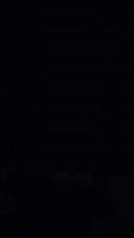 A dark video shows a distant lightning strike illuminating the sky over a silhouetted treeline and a building. A tall pole with a red light on top is visible in the foreground.