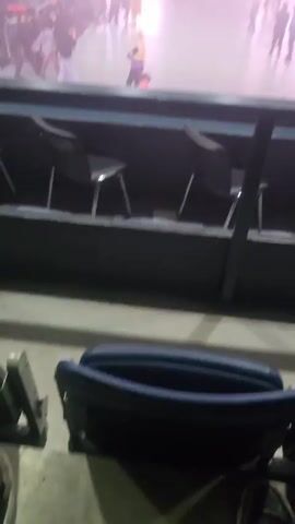 The video shows a crowded indoor event with heavy fog or smoke. People are walking around on a large floor, possibly leaving or arriving. The view is from a higher level behind a barrier and some seating. There are bright lights illuminating the scene. The fog obscures the details of the people and the environment.