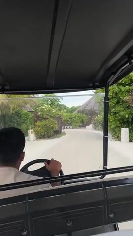 A person is driving a golf cart on a paved path. The person is wearing a light-colored shirt and has short, dark hair. They are holding the steering wheel with both hands. The golf cart has a black roof and clear plastic side panels. The path is lined with lush green trees and bushes, and there are buildings with thatched roofs visible in the background. The ground is sandy. The weather appears to be fair.