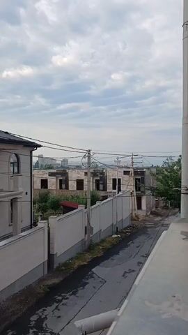 A vertical, slightly tilted shot captures a cloudy sky over a street lined with houses and under-construction buildings. On the left, a beige house with an arched window stands next to a similar, walled property. Across the street, two unfinished buildings with exposed brickwork and window frames are visible, surrounded by greenery. Utility poles and wires crisscross the scene, with a tall concrete pipe on the right edge of the frame. The road is wet and reflects the overcast sky.
