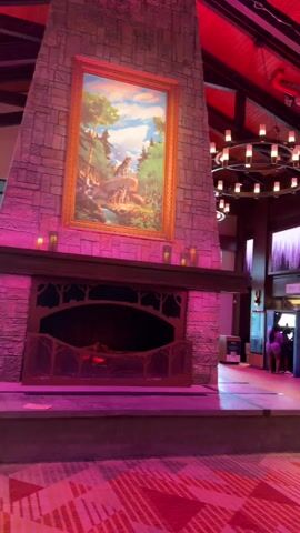 A wide shot of a large, open interior space with a high, beamed ceiling. The space is lit with pink and purple ambient lighting, with prominent circular chandeliers featuring multiple bulbs hanging from the ceiling. To the left is a large stone fireplace with an ornate metal grate and a framed painting of a pastoral scene above it. The floor has a patterned carpet with geometric designs in shades of pink and red. In the background, there are several storefronts, including one labeled "BUCKHORN" and another labeled "EMPORIUM." A large teddy bear mascot is visible standing outside the Buckhorn store. Several people are casually walking through the space. The overall impression is a lobby or common area of a lodge or themed resort.