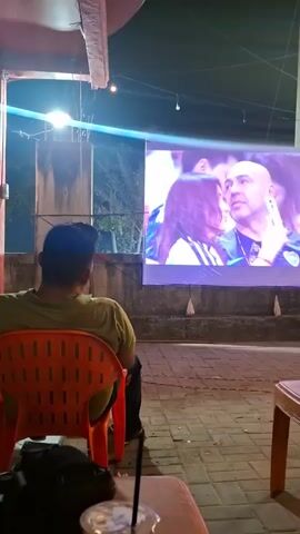 A man sits in an orange plastic chair, facing away from the camera, watching a large outdoor screen. The screen displays a close-up of two people, a man with a shaved head and a woman with long dark hair, both wearing what appears to be sports attire. The man on the screen is looking to his right, while the woman is looking forward. The environment appears to be an outdoor seating area with tiled ground and overhead lighting. A plastic table with a drink is visible in the foreground. The scene is dimly lit, with the screen being the primary source of light.