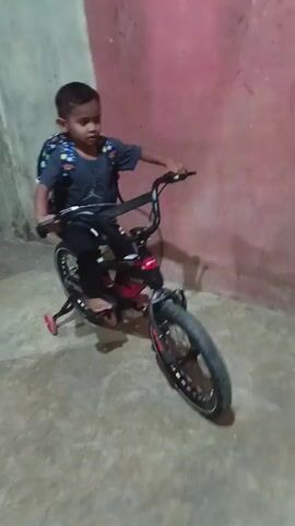 A young child wearing a backpack rides a bicycle. The child is facing forward and appears to be looking at the camera. The bicycle is black and red and has training wheels. The child is in a room with a pink wall and a concrete floor.