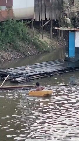 A child is in the water, sitting in a yellow floating object that appears to be a makeshift boat. The child is facing away from the camera and appears to be paddling. In the background, there is a wooden raft with a small structure on it. The water is murky, and the surrounding environment appears to be a developing area with wooden structures and vegetation along the bank.