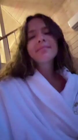 A young woman with long, curly brown hair is wearing a white bathrobe and looking upwards. The background appears to be a wooden room with a sloped ceiling, possibly a cabin or a sauna. The lighting is dim and has a blue hue. The woman's mouth is open as if she is speaking or singing, and her expression is emotional. There is a logo on the right side of her bathrobe.