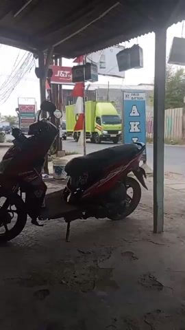 The video shows a roadside scene from under a shelter. In the foreground, two red and white motorcycles are parked. One is closer to the camera, partially obscuring the view. In the background, a road is visible with several vehicles. A large green container truck with a black letter "C" on its side is driving away from the camera. To the left of the container truck, a white truck is approaching. Further down the road, other vehicles and buildings are visible. The environment appears to be an outdoor commercial area with some signs and awnings. The lighting suggests it is daytime.