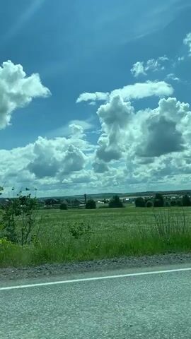 A vehicle travels along a gray asphalt road. On the right side of the road is a large green field. In the distance is a small village. The sky is bright blue with large, puffy white clouds. Trees are visible along the side of the road.