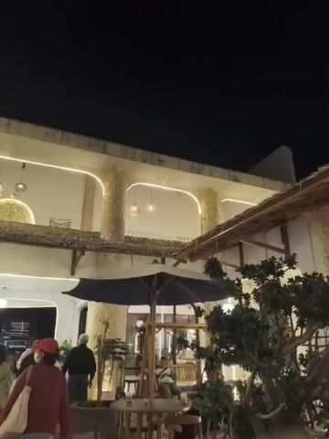 A low-angle outdoor shot at night captures a courtyard with people milling around. The architecture features stone pillars and white walls with arched openings, illuminated by warm-toned string lights that outline the architectural features. In the foreground, a large dark umbrella shades a table with chairs, and a tree with green foliage is visible to the right. Several people are seen walking and standing in the courtyard, some in casual attire. The overall atmosphere appears to be that of a lively outdoor dining or gathering space at night.