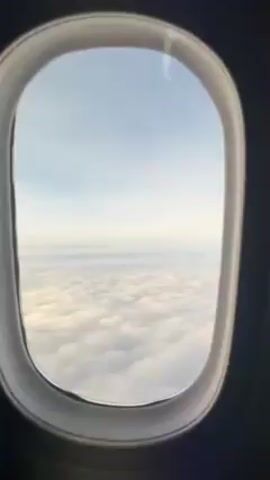 The view from an airplane window shows a dense layer of clouds stretching to the horizon under a pale blue sky. The window's frame is visible, and the overall scene is bright and serene.
