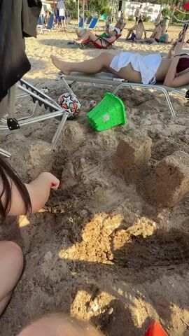 The video captures a beach scene in Nea Potidea, Greece. The foreground focuses on a child's hand digging in the sand with a red toy shovel. Nearby, a partially constructed sandcastle stands, alongside a green plastic bucket and a soccer ball. In the background, several people are relaxing on beach chairs under umbrellas. One person is lying on a lounge chair, covered with a white towel. The sun appears to be shining, casting shadows on the sand, suggesting it's a sunny day, likely in the afternoon. The overall atmosphere appears relaxed and leisurely, typical of a day at the beach. There is no visible text in the video.
