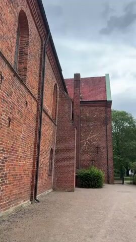 This video pans across a historic building complex with red tile roofs and brick facades. The architecture appears to be traditional, with timber-framed houses visible in the latter half of the clip. The setting is an outdoor, possibly park-like area, with gravel paths, manicured hedges, and mature trees. The sky is overcast with dramatic, grey clouds, suggesting a day with uncertain weather. The overall atmosphere is serene and historical, hinting at a place of cultural or religious significance. There are no people visible in the video, but a dark-colored van is parked in the distance near one of the buildings. The time of day appears to be daytime, given the ambient light. The location is Bassum, Germany.