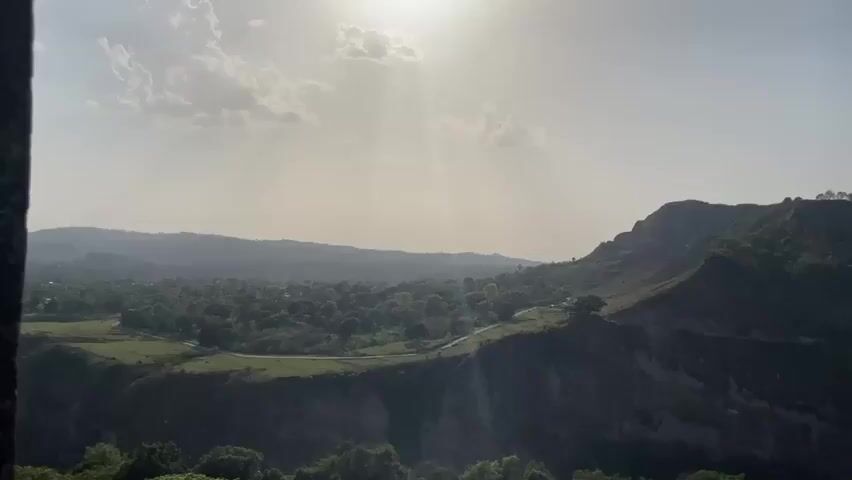 The camera pans across a mountainous landscape. The sun is high in the sky, partially obscured by clouds. The foreground features a steep, forested hillside. In the distance, a valley stretches out, dotted with trees. A winding road snakes along the side of the mountain. As the camera moves, a structure becomes visible atop one of the peaks. Distant mountains appear on the horizon.