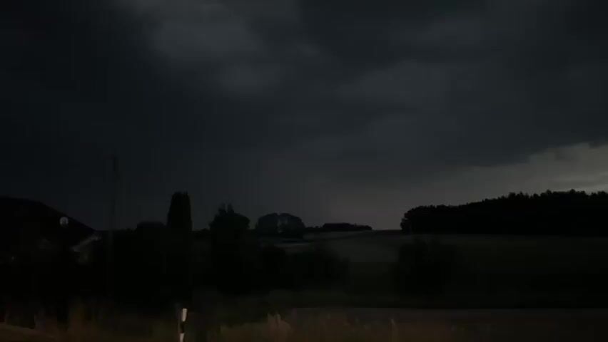 The video shows a dark, stormy sky over a rural landscape. In the foreground, there is a dark road with dry grass. A power pole is visible on the left side of the frame, and a house is partially visible behind it. Trees and fields are spread across the middle ground and background. The sky is filled with dark, heavy clouds, and a bright light, possibly lightning or a distant sunset breaking through the clouds, illuminates the horizon. The video appears to be shot at dusk or during a storm.