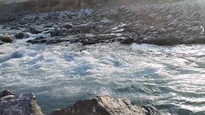 The video shows a river flowing rapidly over rocks. The water is clear and reflects sunlight. The riverbanks are covered in various-sized rocks and pebbles. In the background, distant mountains are visible in the distance. The camera angle shifts, providing different perspectives of the river and its surroundings.