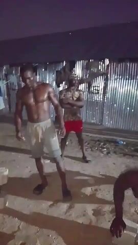 The video shows two men outdoors at night. The man in the foreground is shirtless, wearing light-colored shorts and black sandals. He has a muscular physique and appears to be posing or flexing. He is standing in a sandy or dirt area, with a dumbbell to his left. Behind him, another man is visible, wearing a patterned shirt, red shorts, and black shoes. He is also shirtless and has a muscular build. He appears to be watching the man in the foreground. In the background, there are structures made of corrugated metal and some laundry hanging. The lighting is dim, suggesting it is nighttime.