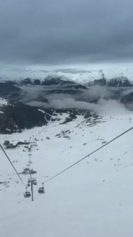 The video shows a snowy mountain landscape from the perspective of someone on a ski lift. In the distance, snow-capped mountains are partially obscured by clouds. The ski lift and its cables are visible, with empty chairs moving along the line. The ground is covered in snow, and there are some buildings and ski runs visible below. The sky is overcast and grey.