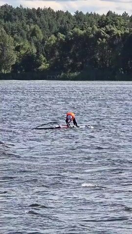 A person wearing an orange life vest and dark clothing is windsurfing on a lake. The windsurfer is bent over the board, appearing to adjust or prepare the sail. The water is choppy with moderate waves, and a line extends from the sail, likely attached to the board. In the background, a tree-lined shore is visible under a cloudy sky.