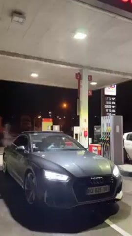 The video shows a gray Audi RS5 coupe at a gas station at night. The car's engine is revving loudly, and its headlights are on. The car is parked under the gas station canopy, and the price display is visible on one of the gas pumps. There is a white car parked next to the Audi. There are no visible people outside the car, but someone appears to be inside. The surrounding environment is dark, but there are streetlights in the background. The camera moves around the car during the revving.
