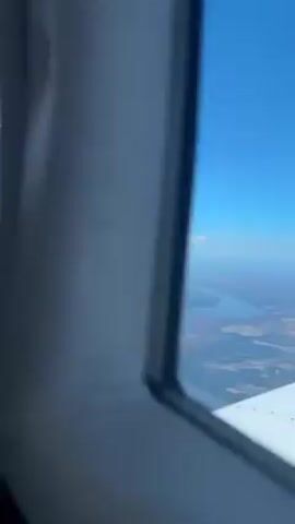 The video shows the interior of a small airplane during flight. A person wearing a cap and Bose headphones is piloting the plane. The plane has a red and black seat in the foreground. The windows show a view of blue sky with scattered clouds. The cockpit instruments are visible. The plane's interior is white with a light-colored headliner. A green tinted sun visor is present.