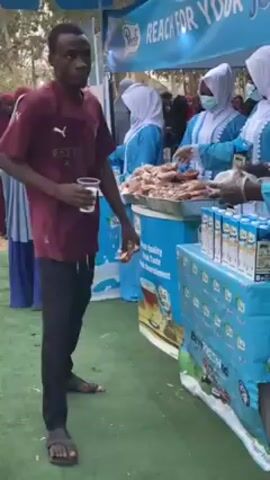 The video shows a crowd of people at an outdoor event. A man in a maroon shirt and black pants holds a plastic cup and walks past a booth with a blue awning that reads "Reach for Your Peak". Several people, mostly women, wearing light blue robes, hijabs, and face masks, stand behind the booth and hand out packages of what appear to be pastries. There is a table with several boxes of what appears to be milk or juice. People walk around the booth, receiving items from the people behind it. There are green mats on the ground and blue and white decorations. Other people are visible in the background, walking around. The overall atmosphere seems festive and organized.