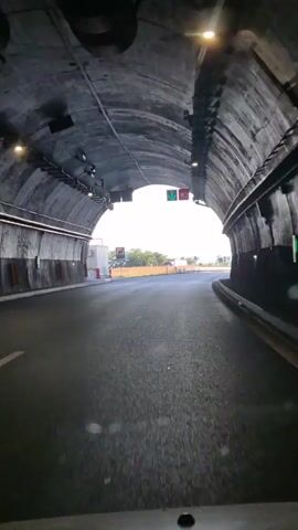 The video shows a first-person perspective of driving through a tunnel and exiting onto an open road. The tunnel is dark with concrete walls and ceiling, illuminated by artificial lights. As the vehicle approaches the tunnel exit, the bright sunlight becomes visible. Outside the tunnel, the road curves to the right, with green traffic lights indicating the lane. The surrounding environment appears to be rural, with hills and mountains visible in the distance. The sky is clear and blue, and the lighting suggests it is daytime. There are no people visible in the video.