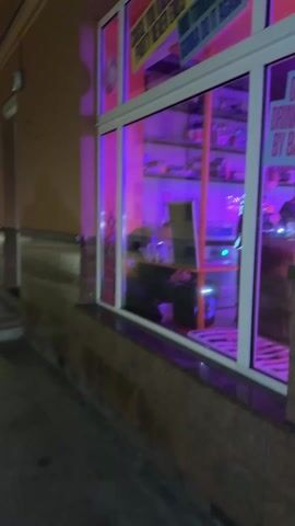 The video shows a night scene of a narrow, cobblestone street lined with buildings on both sides. The buildings are mostly two to three stories tall and appear to be of an older architectural style with lit windows. String lights illuminate the street on the right-hand side, near what looks like outdoor cafe seating. A person is visible standing near the cafe. In the distance, the street continues, with more buildings visible, some illuminated with colored lights. The sky is dark and the overall lighting is low.  The camera moves slightly, giving a sense of a slow walk or scan of the area.