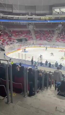 The video shows a hockey game in progress at an indoor arena. The arena is filled with spectators, mostly seated in red chairs, although some are standing. Two cheerleaders with blue pom-poms are visible in the foreground. The hockey players, wearing teal and dark colored uniforms, are actively skating on the ice. Several officials are present on the sidelines. A large screen hangs above the ice rink, displaying the game score, time and possibly advertisements. Banners and sponsor logos are visible around the perimeter of the rink. The camera is positioned in the stands, capturing a wide view of the game and the audience.
