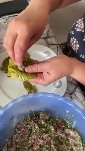 A close-up video shows a pair of hands preparing a dish. The hands are carefully wrapping a mixture of ground meat and herbs within a grape leaf. The mixture is in a blue bowl in the foreground, and several other grape leaves are on a white plate in the background. The hands are using their fingers to tuck the edges of the grape leaf around the filling, creating a neat package. The scene appears to be taking place in a kitchen, with a patterned countertop visible.