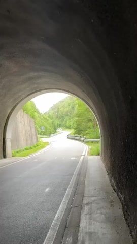 The video shows a view from inside a tunnel looking out towards a curved asphalt road that winds its way up a tree-covered hill. The tunnel has a concrete ceiling and walls. To the left of the road, there is a concrete retaining wall covered in greenery. To the right, there is a sidewalk and a metal guardrail. The road is wet, suggesting recent rain. The sky is overcast. There are no people or vehicles visible in the video. The overall impression is one of a quiet, natural setting.