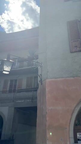 The video shows a collection of clips of a building, likely a museum, in a sunny outdoor environment. The first clip shows a close-up of the building’s architecture, featuring a window with brown shutters and an arched doorway below. The second clip pans across the exterior of a building with yellow walls and many windows, framed against a bright blue sky with a few clouds. The third clip shows a sign that reads “MUSEE DU VIEUX-COPPET” (Museum of Old Coppet), with a tree overhead and tables and chairs in the background. The final clip shows a street scene with a building on a raised platform, with multiple flags and signs around it. People are walking on the street in the background.