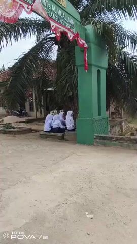 A group of four young people, appearing to be students wearing white uniforms and hijabs, are seated on a low concrete wall near a green archway. They are facing away from the camera, with their heads bowed, possibly engaged in conversation or prayer. Behind them, a building with a tiled roof is visible, along with palm trees and other vegetation. The ground in the foreground is dirt and gravel. The archway has text written on it and is decorated with red and white banners. The overall scene suggests an outdoor area of a school or religious institution.