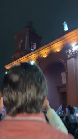 A night-time video shows a crowd of people gathered in front of a building with columns and illuminated windows. Many individuals are holding up their phones, presumably to record or take pictures. Several people in the crowd are waving Argentine flags. The atmosphere appears celebratory.
