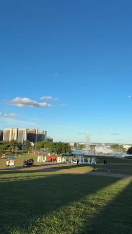 The video captures a daytime scene in a park, likely in Brasília, Brazil, given the prominent "EU BRASILIA" sign. The sky is a clear, vibrant blue with a few wispy clouds, suggesting pleasant weather. A large fountain is the central focus of the background, with water jetting high into the air and a significant amount of mist and spray surrounding its base. In the distance, several modern buildings and structures are visible, hinting at an urban environment.

In the foreground, a lush green lawn dominates the lower half of the frame, with shadows cast from unseen trees, indicating the sun is at a particular angle, likely late afternoon or early morning. The large white letters spelling out "EU BRASILIA" are positioned on the lawn, with people interacting around them. Some individuals are posing for photos, while others are walking or observing the fountain. A couple of food carts are also present, suggesting a recreational area where visitors can purchase refreshments. The overall mood appears relaxed and enjoyable, with people taking advantage of the outdoor space and the iconic landmark. The presence of the moon in the sky, though faint, is a notable detail.