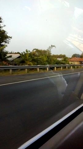 The video shows a view from inside a vehicle, looking out a window. The window has a smudge or water stain on it. To the left, through the window, is a bright, overcast sky and a landscape with trees, some of which appear to be banana trees, and buildings. A road is visible running parallel to the vehicle, and to the right is the interior of the vehicle, including a dark-colored seat or paneling. The perspective suggests the vehicle is in motion.