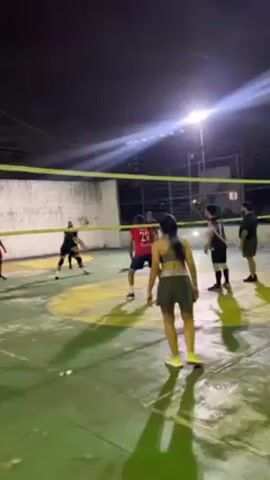 The video shows a group of people playing volleyball on an outdoor court at night. The court has a green surface and is surrounded by a white brick wall and a fence. A volleyball net is stretched across the center of the court.

There are about 10 players visible, wearing athletic attire. Most are standing on the court, poised to play. The lighting is provided by bright overhead lamps that cast a strong glare.

Throughout the video, the volleyball is actively being played. The players are seen bumping, setting, and spiking the ball. They react to the movements of the ball and each other, displaying teamwork and engagement in the game.